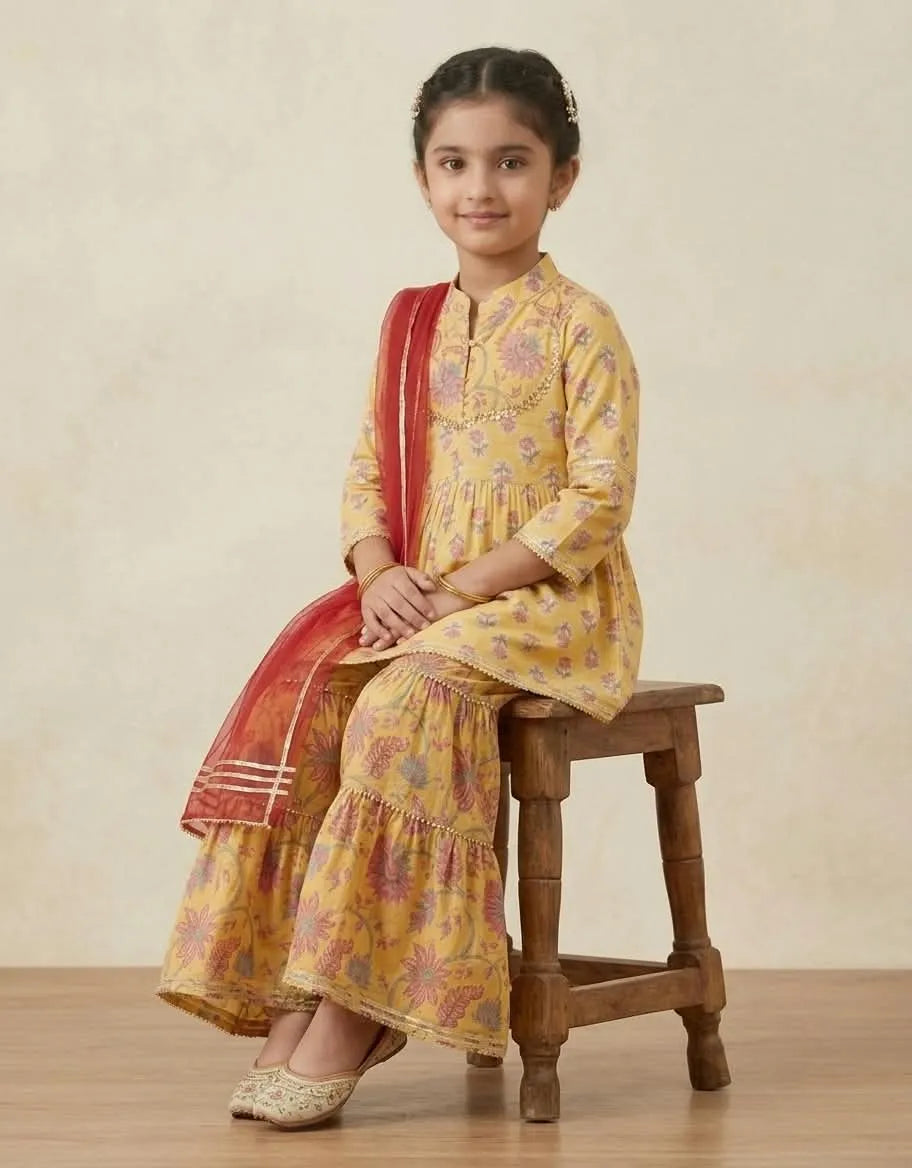 A young girl wearing a yellow floral printed salwar kameez suit with a red dupatta, sitting on a wooden stool against a neutral beige background.