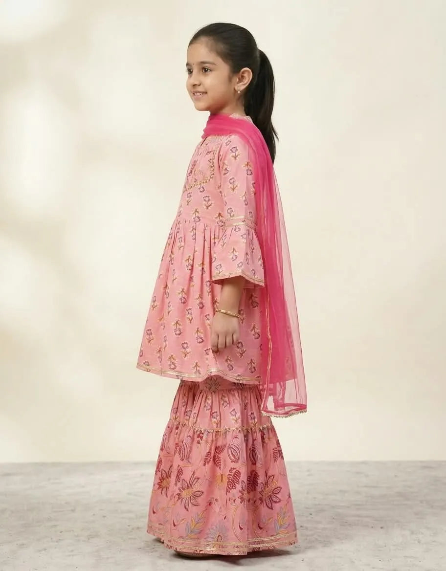 A young girl with her hair in a ponytail is shown from the side, wearing a pink tiered salwar kameez suit with floral prints and a bright pink dupatta, set against a neutral beige studio background.