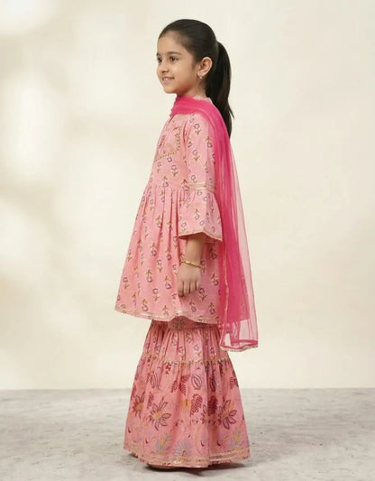 A young girl with her hair in a ponytail is shown from the side, wearing a pink tiered salwar kameez suit with floral prints and a bright pink dupatta, set against a neutral beige studio background.