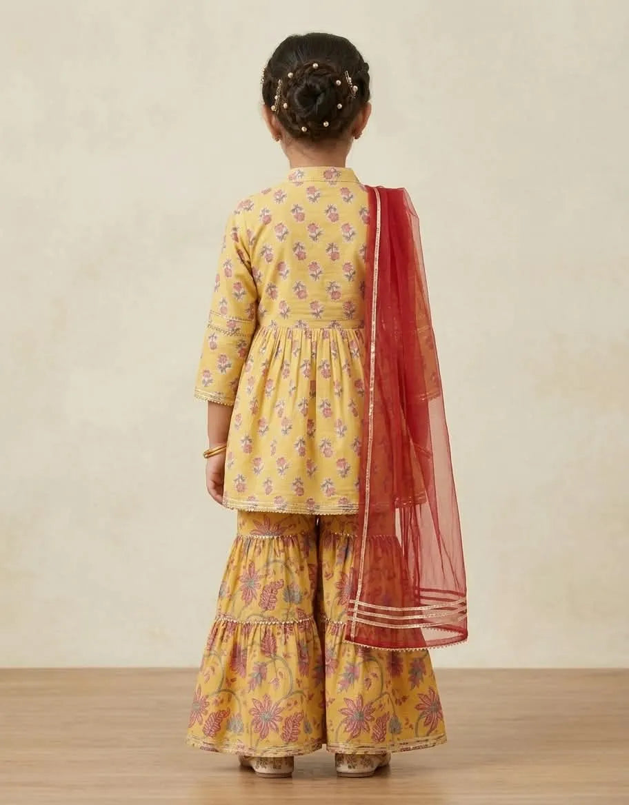 A young girl viewed from behind wearing a yellow printed kurta and flared pants with a red dupatta, standing against a neutral beige wall.