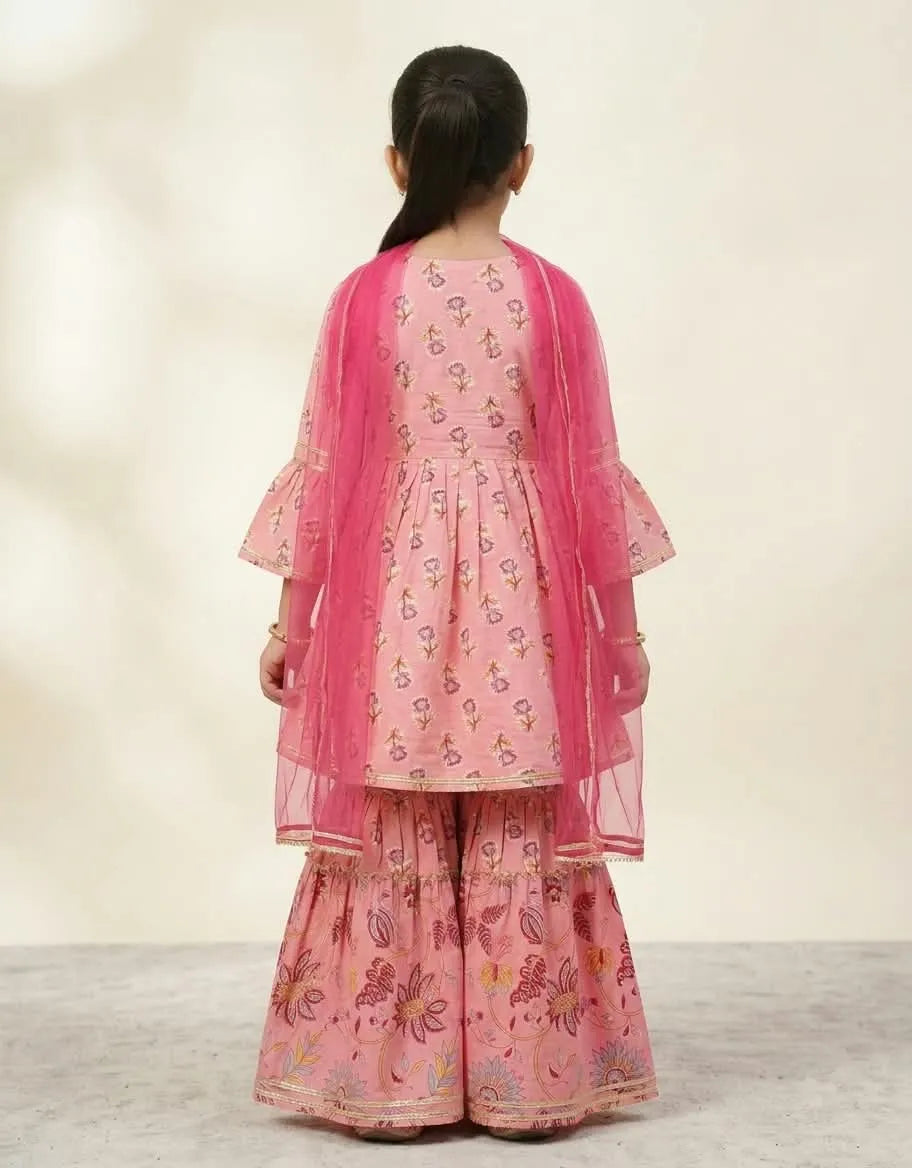 A young girl viewed from behind wearing a pink traditional outfit with a sheer pink dupatta and floral printed fabric, standing against a plain light beige studio background.