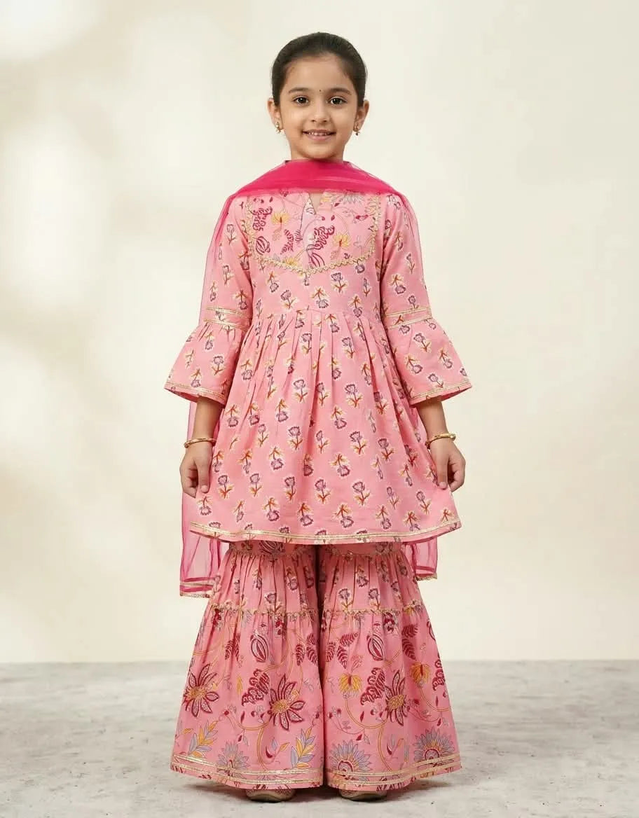 A young girl modeling a pink printed traditional outfit with a matching dupatta, featuring a kurta with bell sleeves and wide flared pants, set against a neutral studio background.