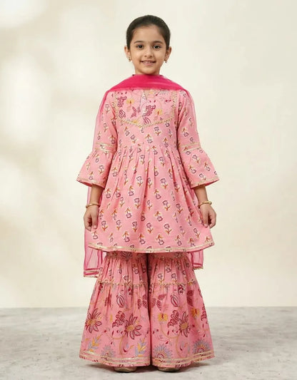 A young girl modeling a pink printed traditional outfit with a matching dupatta, featuring a kurta with bell sleeves and wide flared pants, set against a neutral studio background.
