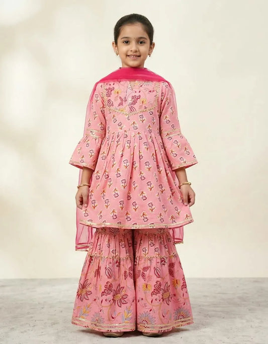 A young girl modeling a pink printed traditional outfit with a matching dupatta, featuring a kurta with bell sleeves and wide flared pants, set against a neutral studio background.
