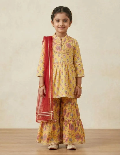 A young girl modeling a yellow floral print kurta and flared pants set with a red dupatta, standing against a neutral beige background.