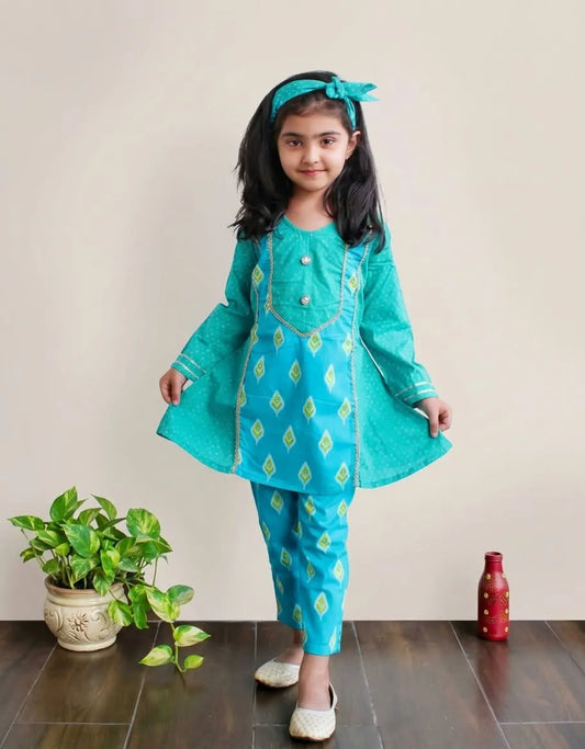 A young girl modeling a turquoise blue traditional Indian outfit with yellow diamond patterns, consisting of a long kurta, matching pants, and a headband, standing in a room with a potted plant and a red bottle.