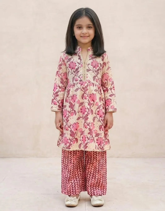 A young girl stands against a plain beige wall wearing a traditional Indian outfit consisting of a beige kurta with pink floral patterns and matching red patterned pants.