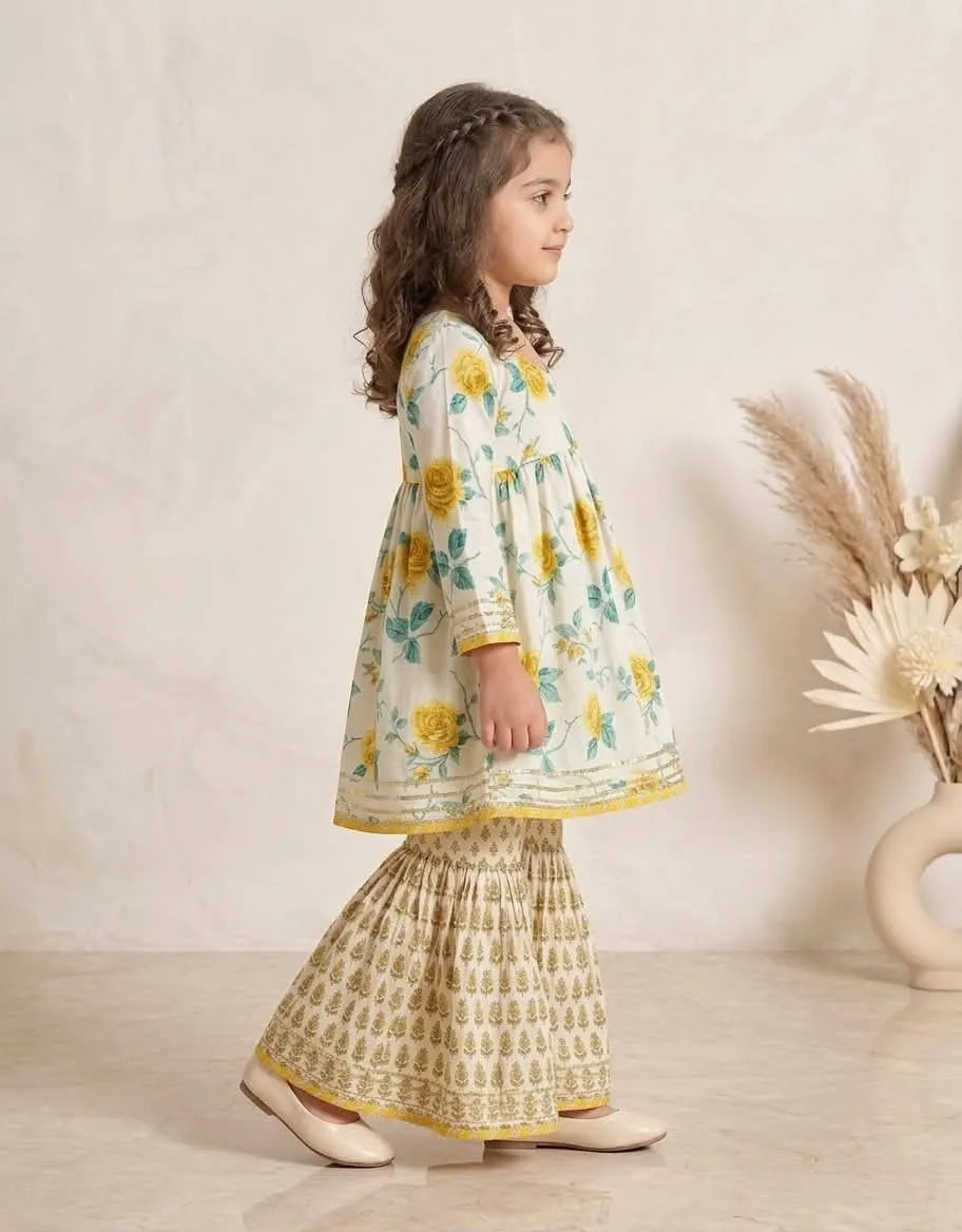 A young girl with long brown hair is shown from the side, wearing a traditional Indian outfit consisting of a cream-colored kurta with yellow and green floral patterns, paired with a matching printed flared skirt. She is standing in a studio setting with a textured beige wall and a decorative vase of dried grass on the right.