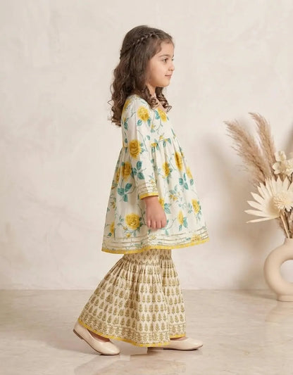 A young girl with long brown hair is shown from the side, wearing a traditional Indian outfit consisting of a cream-colored kurta with yellow and green floral patterns, paired with a matching printed flared skirt. She is standing in a studio setting with a textured beige wall and a decorative vase of dried grass on the right.