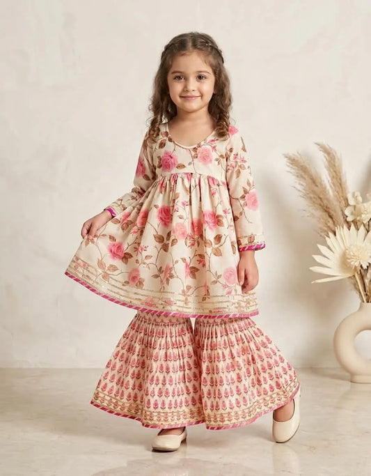 A young girl modeling a traditional Indian three-piece salwar kameez set. The set features a beige kameez top with pink floral prints, matching pink and beige patterned salwar pants, and a beige border skirt.