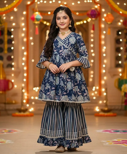 A woman modeling a navy blue and white printed traditional Indian outfit with wide-leg striped pants, standing in a decorated indoor setting with warm string lights.