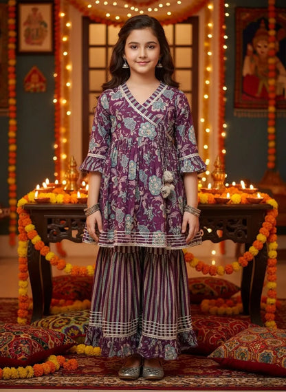 A young girl stands in a decorated indoor setting wearing a purple and white printed traditional tunic and striped palazzo pants. The background features warm string lights, marigold garlands, and lit candles on a dark wooden table.