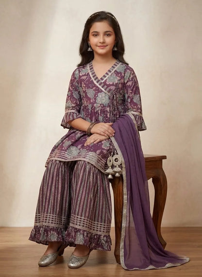 A young girl models a traditional Indian salwar kameez suit in purple and silver, sitting on a wooden stool against a neutral beige background.