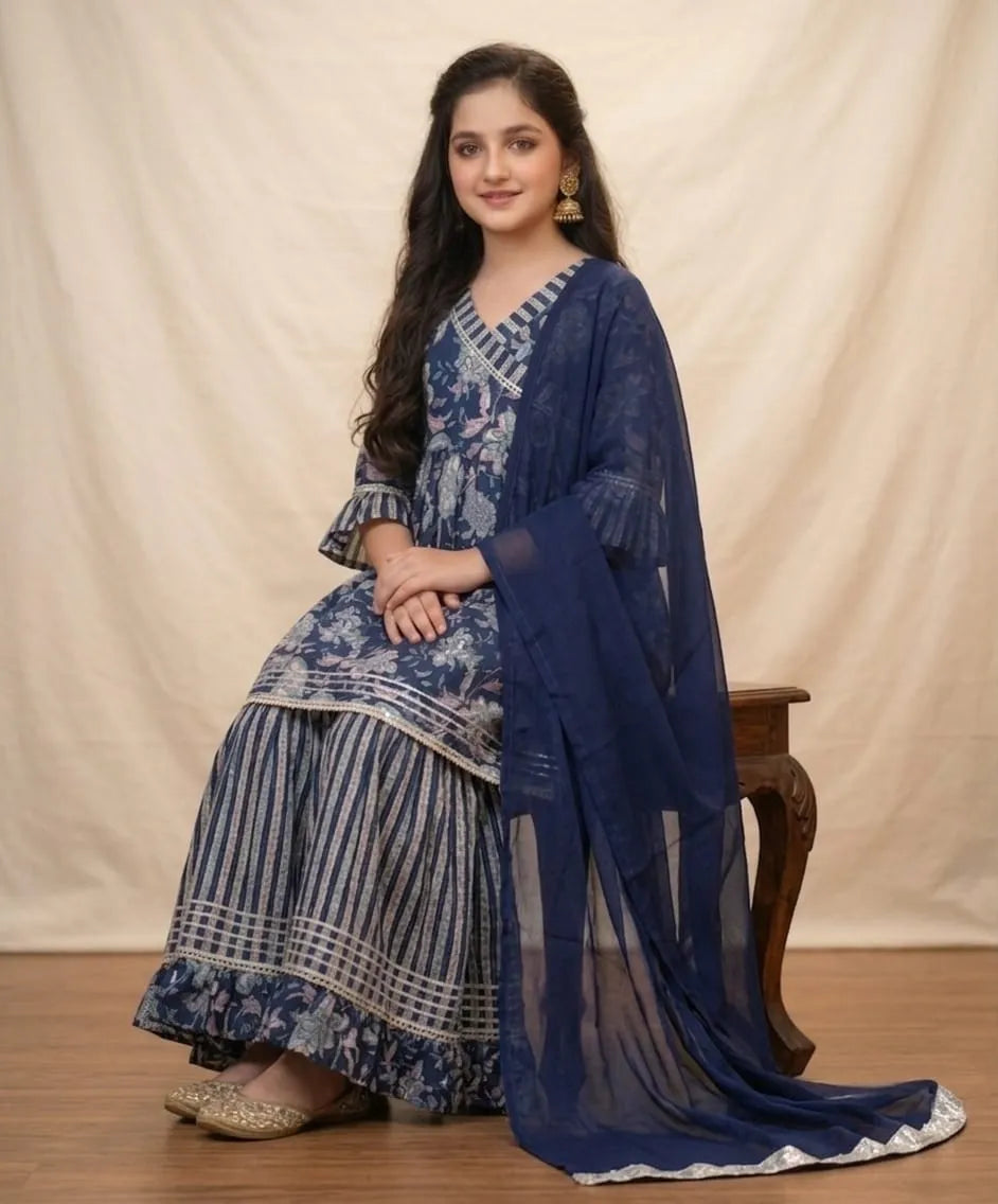 A woman models a traditional Indian salwar kameez suit in navy blue and white, featuring a printed kurta, striped palazzo pants, and a matching sheer dupatta, seated on a wooden stool against a cream-colored backdrop.