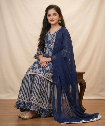 A woman models a traditional Indian salwar kameez suit in navy blue and white, featuring a printed kurta, striped palazzo pants, and a matching sheer dupatta, seated on a wooden stool against a cream-colored backdrop.