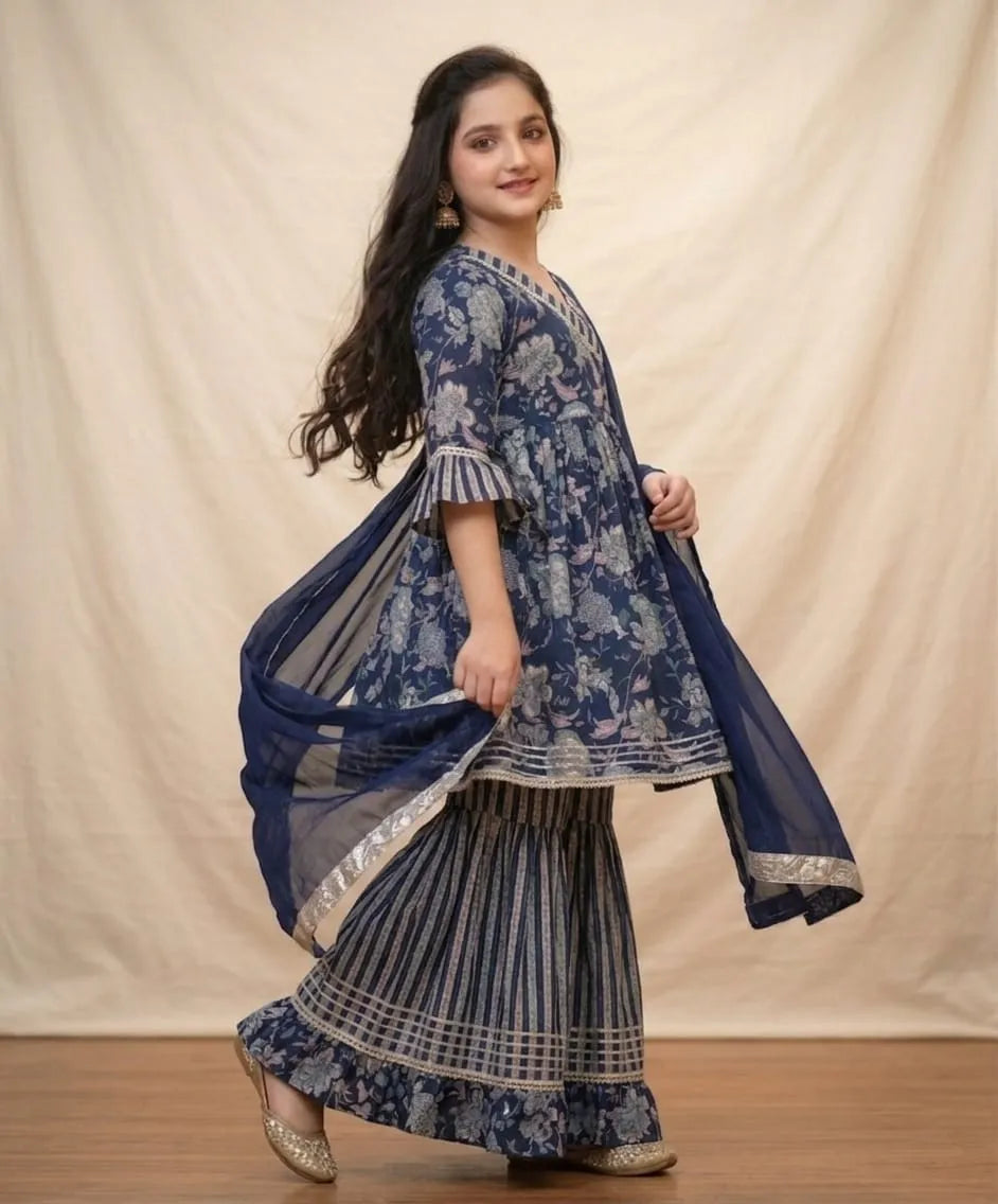 A woman modeling a traditional Indian outfit consisting of a navy blue floral printed kurta, pleated flared pants, and a matching sheer dupatta, set against a neutral draped backdrop.