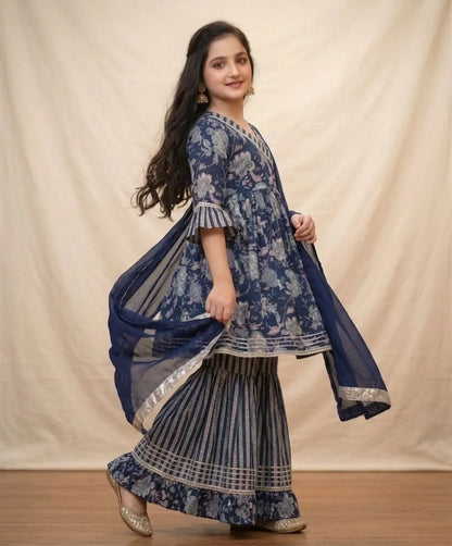 A woman modeling a traditional Indian outfit consisting of a navy blue floral printed kurta, pleated flared pants, and a matching sheer dupatta, set against a neutral draped backdrop.