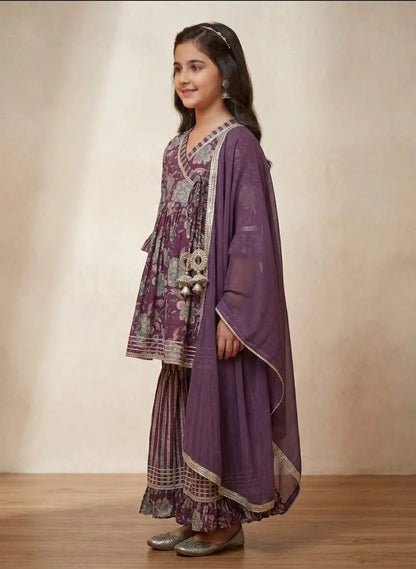 A young girl modeling a traditional purple salwar kameez suit with intricate silver and white embroidery, paired with a matching sheer purple dupatta, set against a neutral beige studio backdrop.