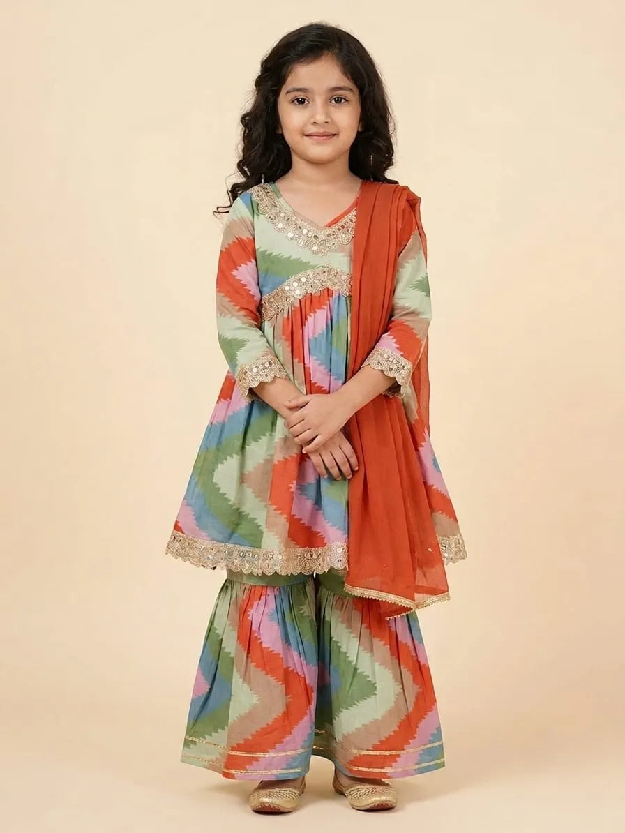 A young girl modeling a traditional Indian outfit consisting of a multicolor zigzag-patterned kurta and sharara pants with a rust-orange dupatta, set against a solid beige background.