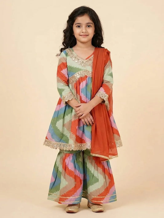 A young girl modeling a traditional Indian outfit consisting of a multicolor zigzag-patterned kurta and sharara pants with a rust-orange dupatta, set against a solid beige background.