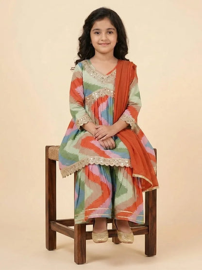 A young girl sits on a wooden stool wearing a colorful patterned traditional outfit with a solid orange dupatta, set against a plain beige studio background.