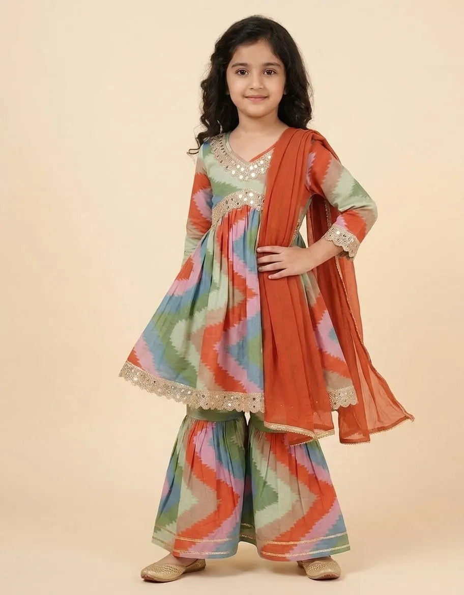 A young girl modeling a traditional Indian outfit consisting of a multicolor zigzag-patterned kurta and flared pants with a rust-orange dupatta, set against a solid beige background.