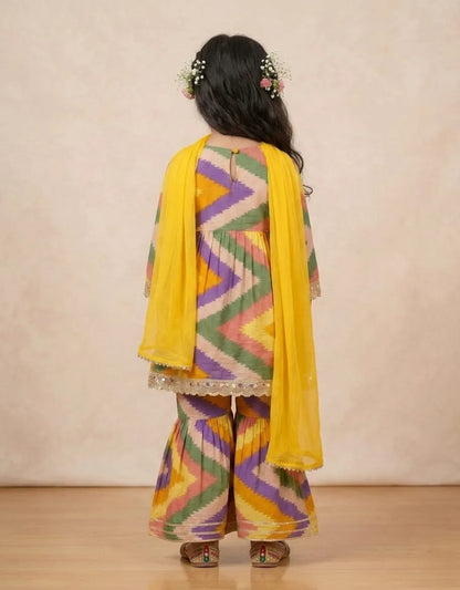 A child model shown from the back wearing a traditional Indian outfit with a multicolor zigzag patterned kurta and wide-leg pants, paired with a bright yellow dupatta, against a neutral beige background.