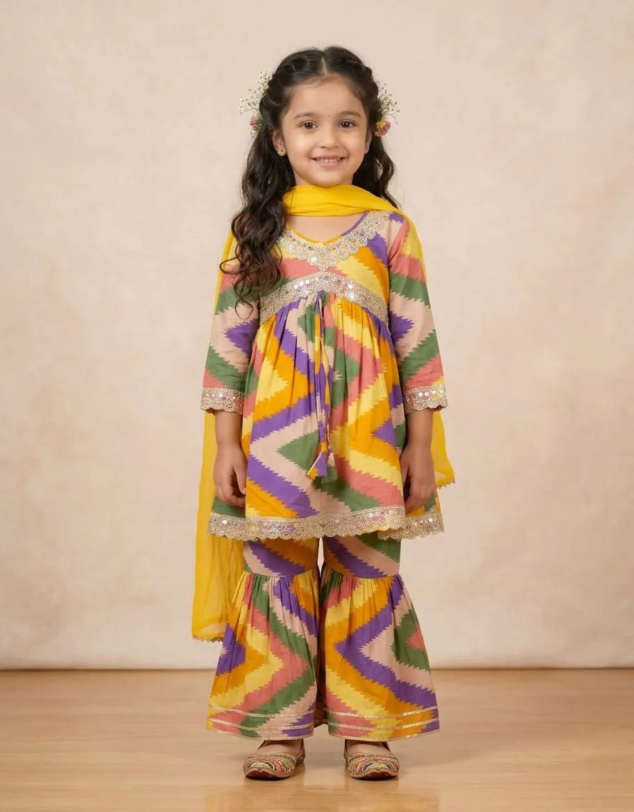 A young girl modeling a vibrant, multicolored traditional Indian outfit with a yellow dupatta, featuring a chevron pattern in yellow, green, purple, pink, and orange, set against a neutral beige studio backdrop.