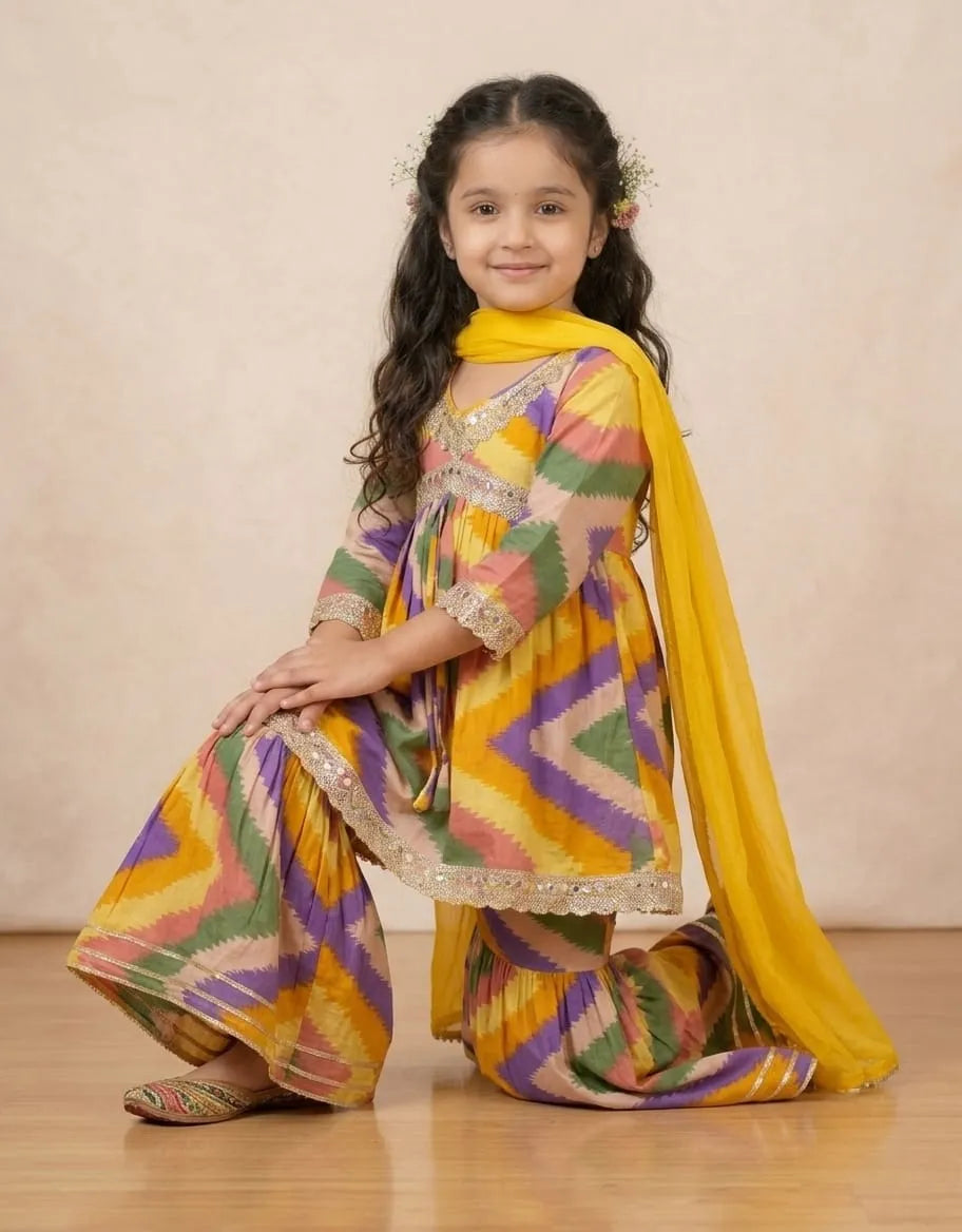 A young girl kneeling and smiling while wearing a colorful chevron-patterned traditional outfit with a bright yellow dupatta, set against a neutral beige studio backdrop.