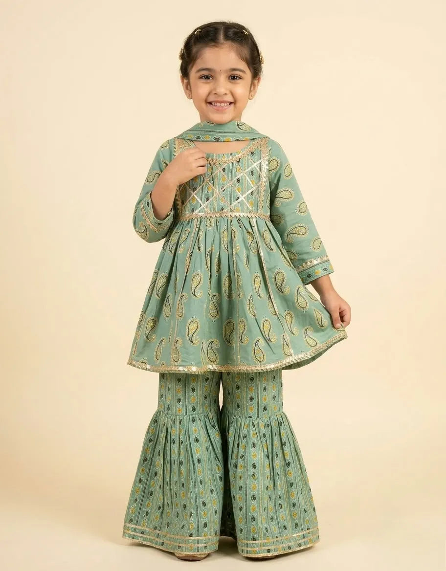 A young girl modeling a mint green traditional outfit with gold paisley patterns and gold trim, consisting of a long-sleeve kurta and matching flared pants, set against a solid beige background.