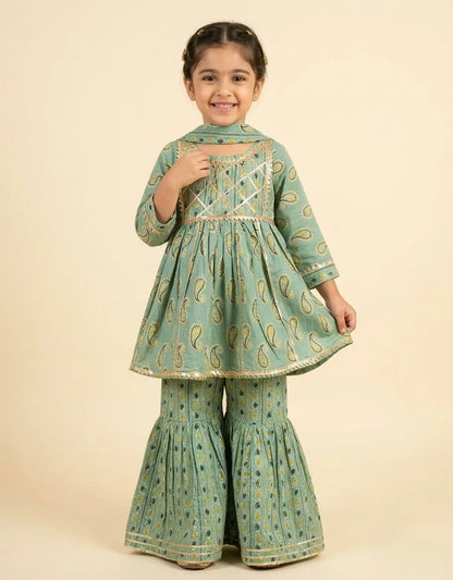 A young girl modeling a mint green traditional outfit with gold paisley patterns and gold trim, consisting of a long-sleeve kurta and matching flared pants, set against a solid beige background.