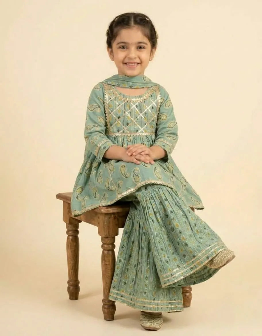 A young girl sitting on a wooden stool wearing a mint green traditional Indian outfit with gold embroidery and a matching dupatta, set against a plain beige background.