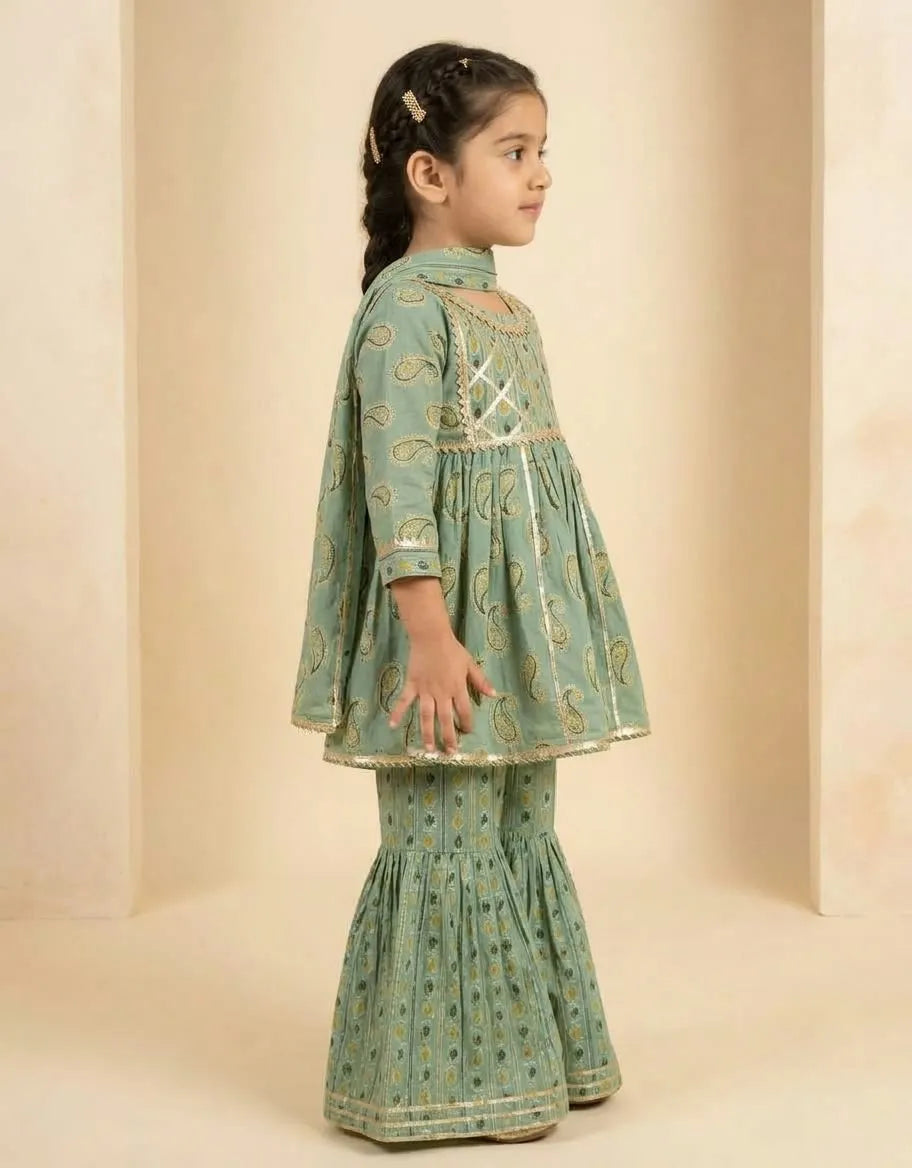 A young girl modeling a sage green traditional outfit with gold paisley patterns, standing in profile against a beige background.