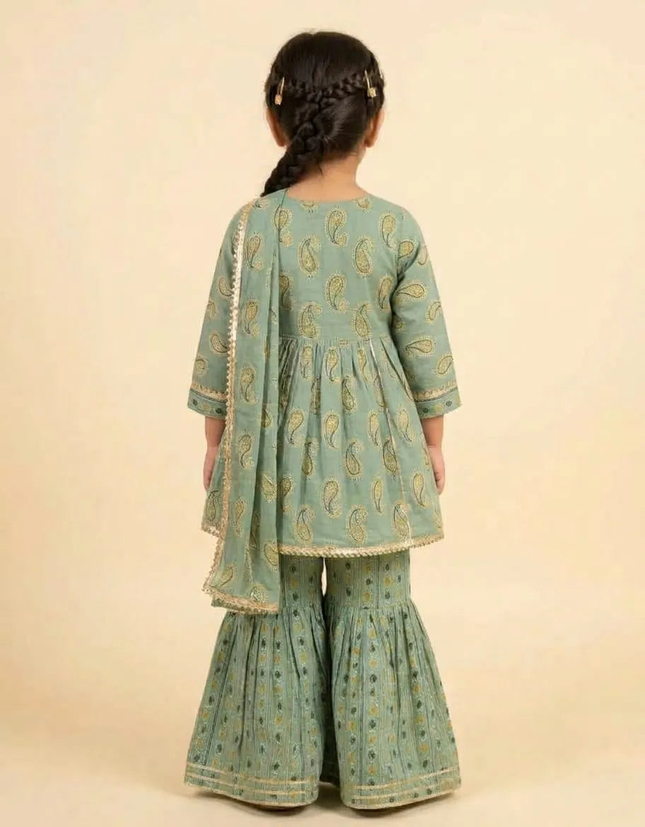 A young girl viewed from behind wearing a sage green traditional outfit with gold paisley patterns and a matching dupatta, set against a solid beige background.