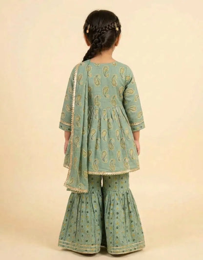 A young girl viewed from behind wearing a sage green traditional outfit with gold paisley patterns and a matching dupatta, set against a solid beige background.
