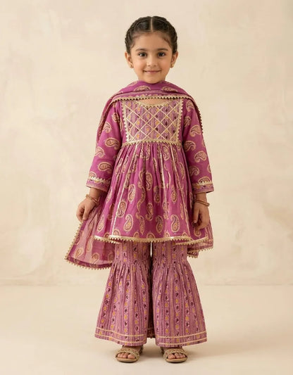 A young girl modeling a traditional Indian outfit consisting of a magenta pink kurta with gold embroidery, matching flared pants, and a dupatta, set against a plain beige background.