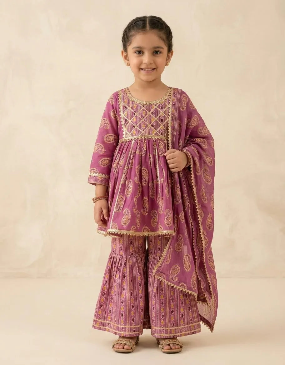 A young girl modeling a traditional Indian outfit consisting of a magenta pink kurta with gold paisley patterns, matching flared pants, and a coordinating dupatta, set against a warm beige studio backdrop.