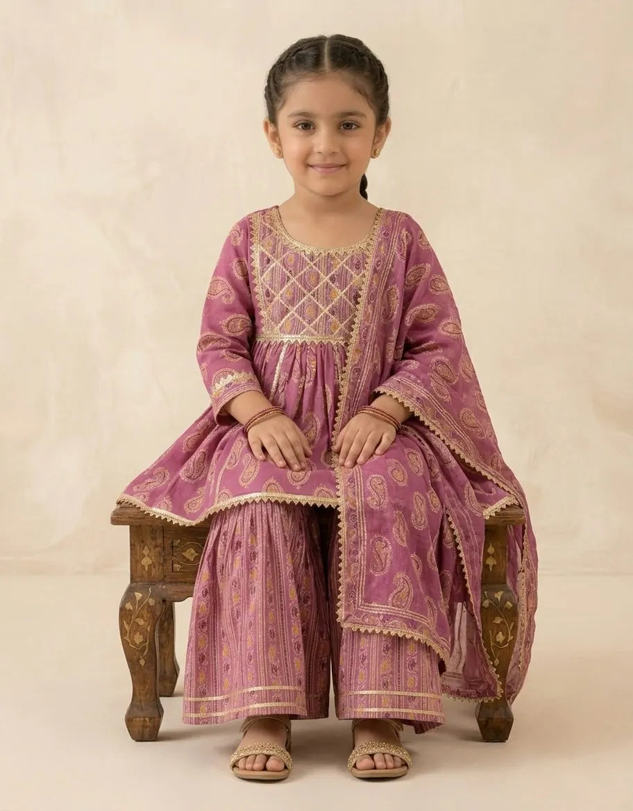 A young girl sits on a carved wooden stool wearing a traditional Indian salwar kameez suit in dusty pink with gold paisley patterns and embroidery. The image is shot against a neutral beige studio backdrop.