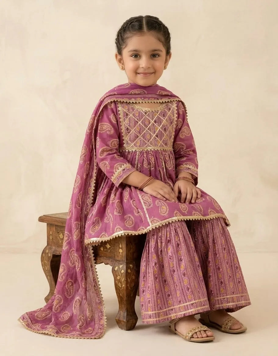 A young girl wearing a traditional Indian outfit in dusty pink and gold, sitting on a small wooden stool against a warm beige background.