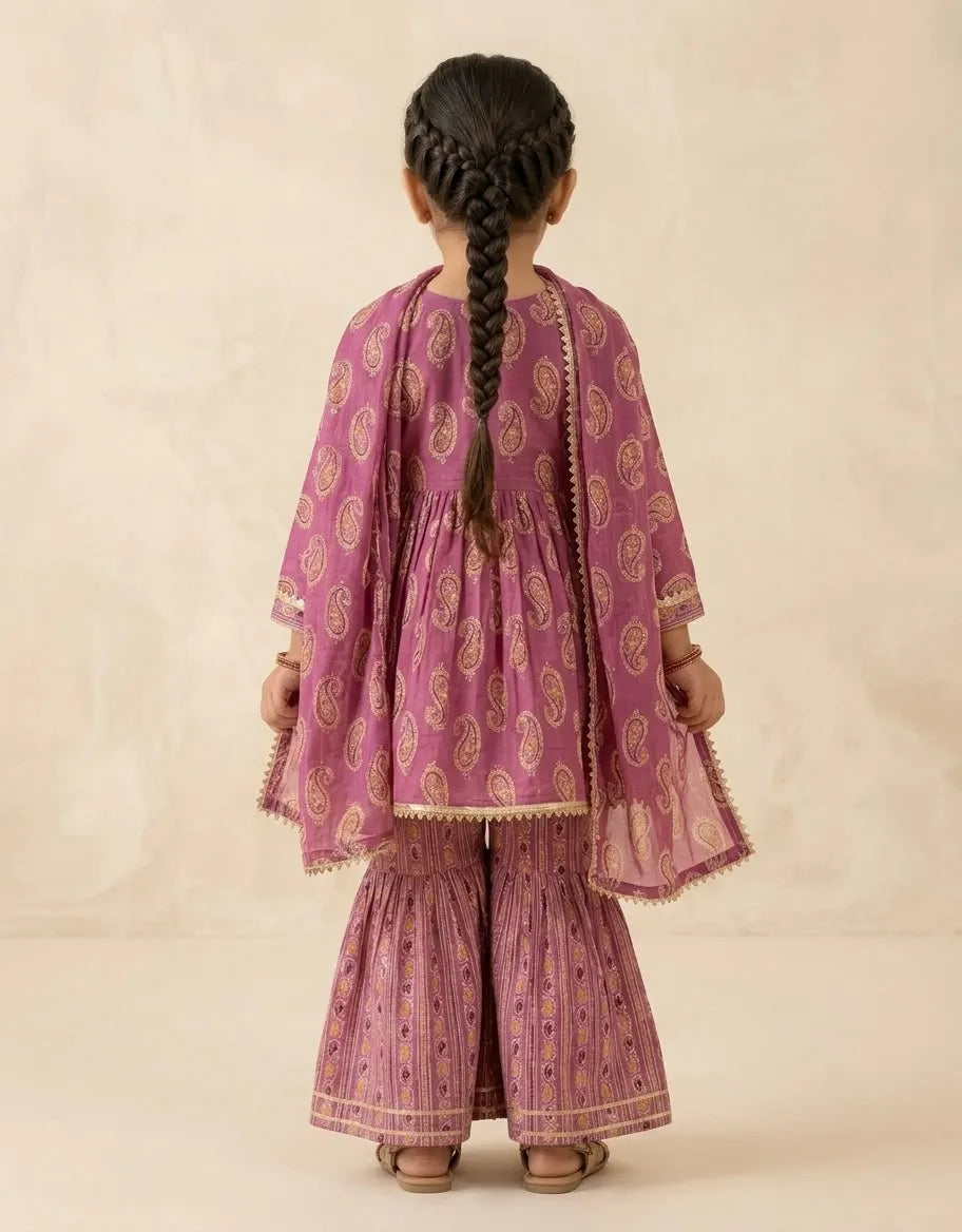 A young girl viewed from behind wearing a traditional Indian outfit consisting of a dusty purple kurta and matching wide-leg pants with gold paisley patterns, set against a neutral beige background.