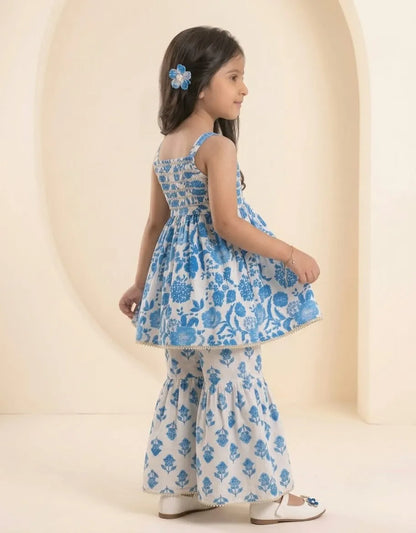 A young girl wearing a blue and white printed sleeveless kurta with matching flared pants, standing in profile against a warm beige arched studio backdrop.