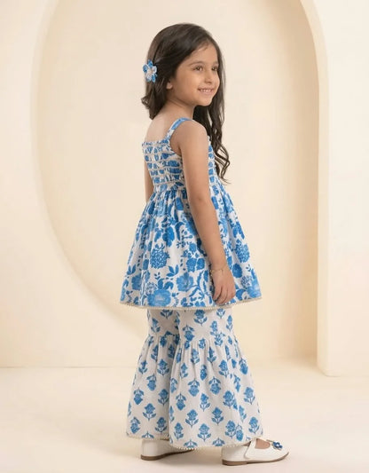 A young girl with long dark hair is shown from the back wearing a blue and white printed sleeveless dress with matching flared pants. She is standing in a studio with a warm beige background featuring arched architectural details.