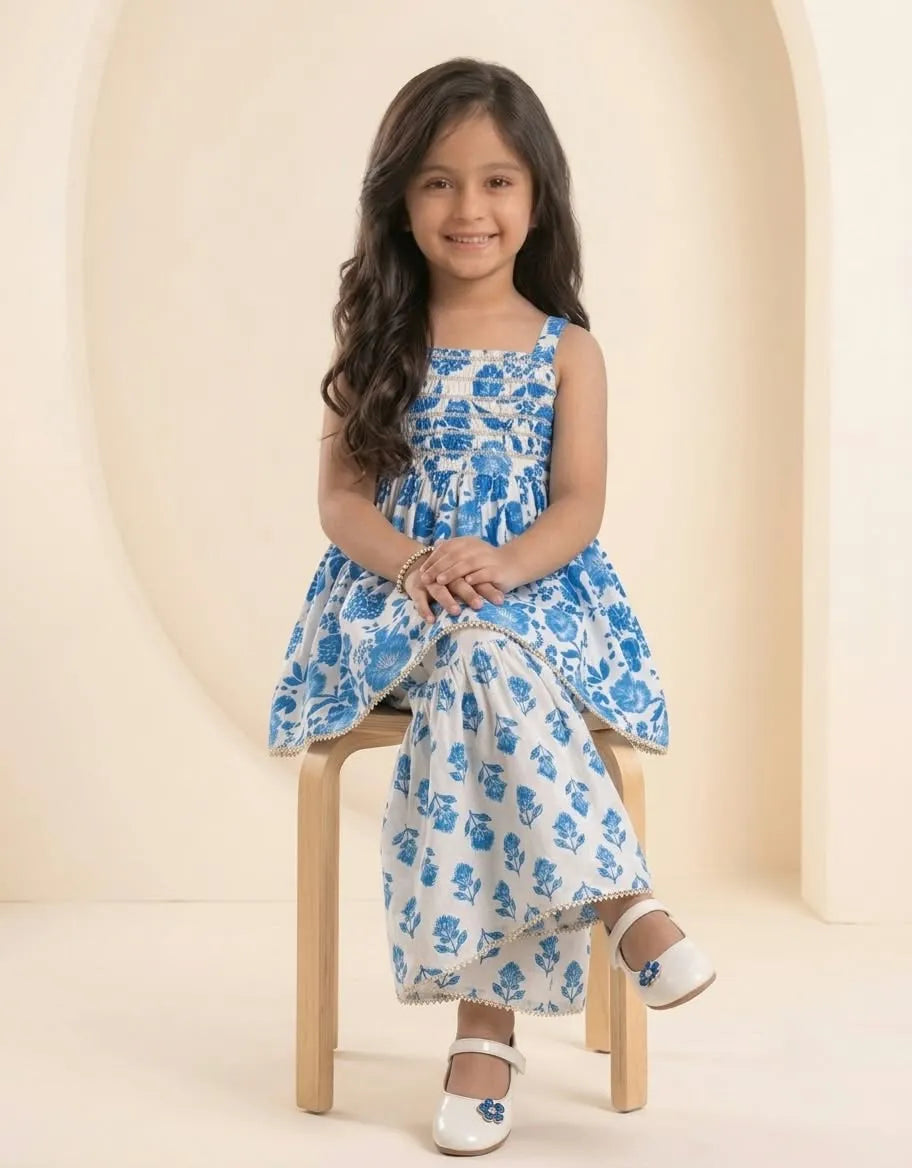 A young girl with long dark hair sits on a small wooden stool, wearing a sleeveless blue and white floral print dress with matching white shoes.