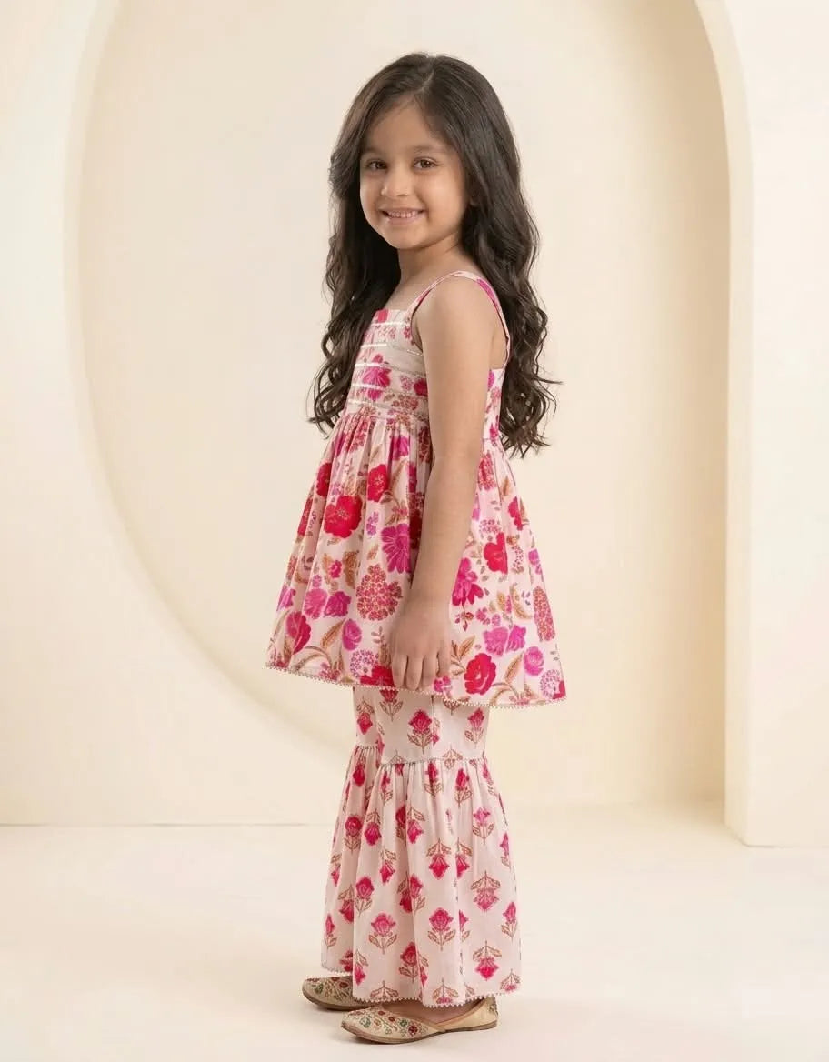 A young girl with long dark hair stands in profile wearing a sleeveless tiered dress with a pink and red floral print on a light beige background. She is standing in front of a warm cream-colored studio backdrop with an arched opening.