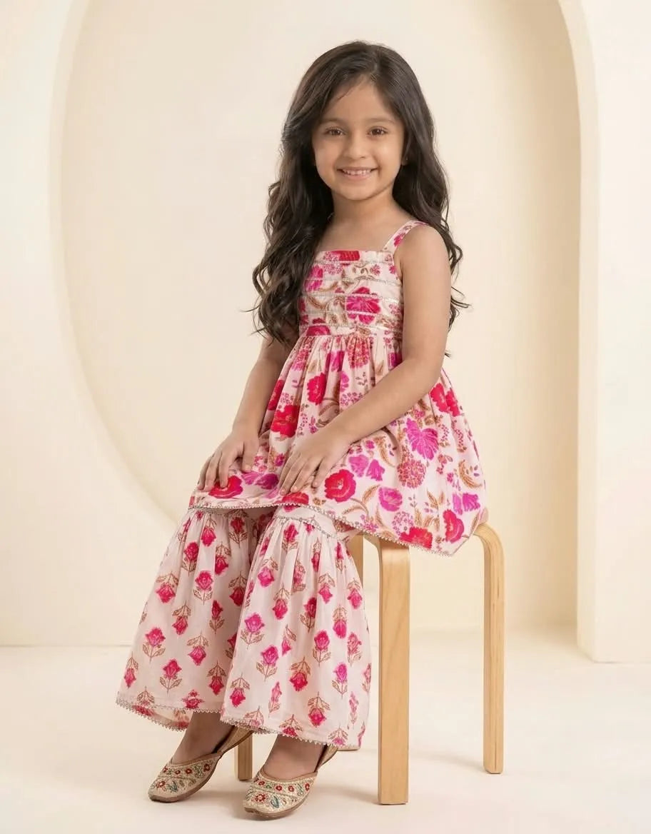 A young girl with long dark hair sits on a wooden stool, wearing a sleeveless tiered dress with a pink and red floral print on a cream background. The dress features thin shoulder straps and a gathered waist.