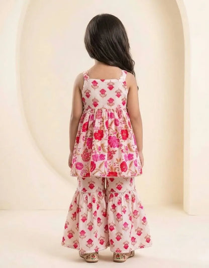 A young girl viewed from behind wearing a two-piece floral print outfit with a sleeveless peplum-style top and matching flared pants, set against a warm cream-colored arched studio backdrop.