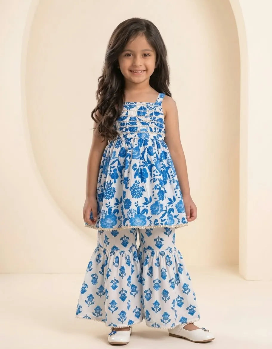A young girl with long dark hair stands in a studio wearing a sleeveless white and blue floral printed tunic top with matching wide-leg pants. The background is a warm beige with an arched architectural shape.