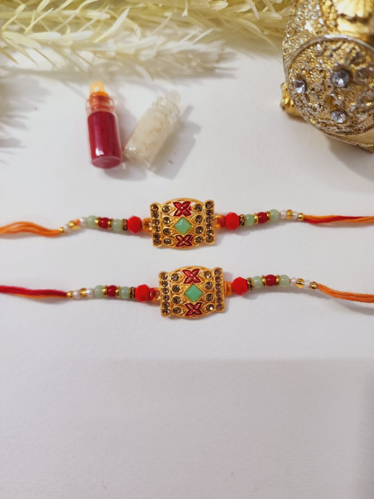 Two designer rakhis with golden embellishments and multicolored beads, presented alongside a small red bottle and decorative golden elements.