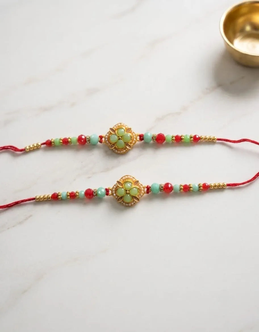 Two red string rakhi bracelets with gold-plated floral center ornaments and green and red beads, displayed on a white marble surface with a small gold bowl in the top right corner.