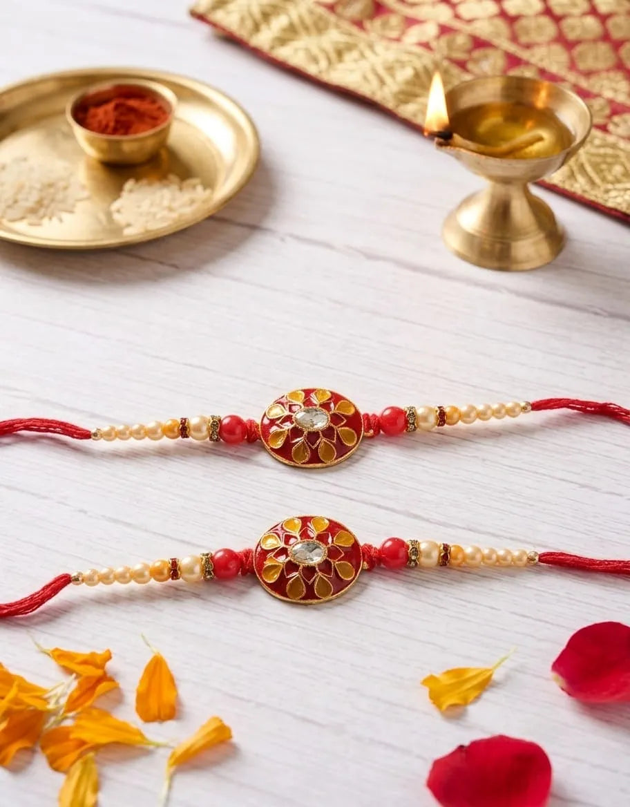 Two traditional Indian Rakhi beaded wristlets with ornate gold and red circular medallions, displayed on a white wooden surface with festive props including a brass diya, a plate with rice and red powder, and flower petals.